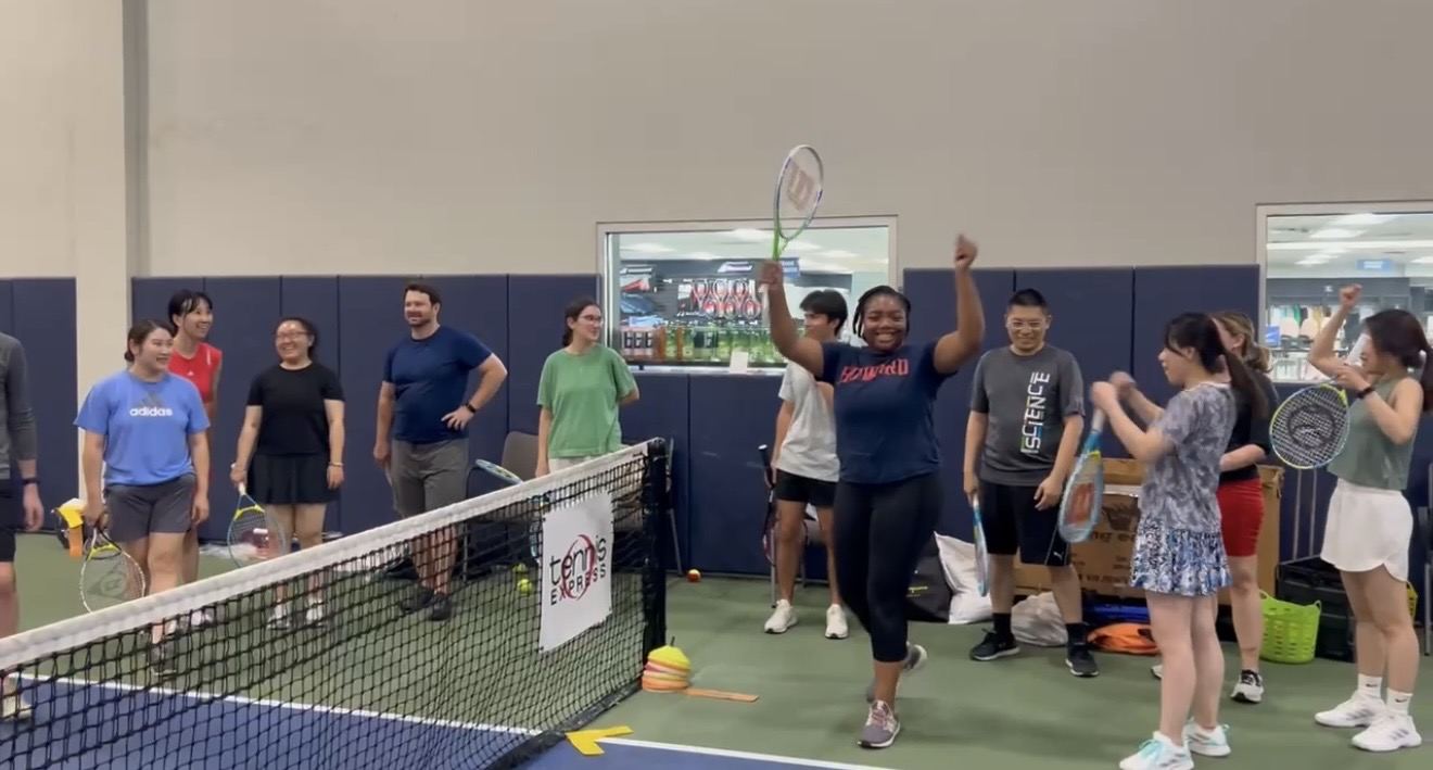 An adult beginner raising her racquet and both arms after winning a point, with the rest of her group cheering behind the net