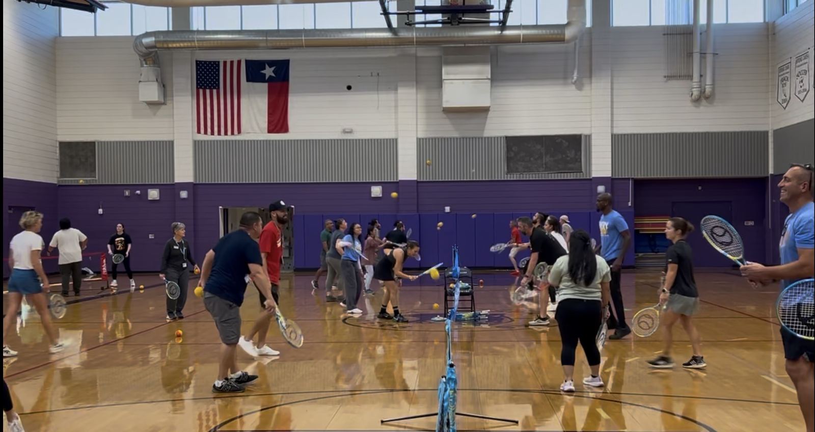 About twenty-five adults spread across portable nets in a school gym, rallying with orange balls