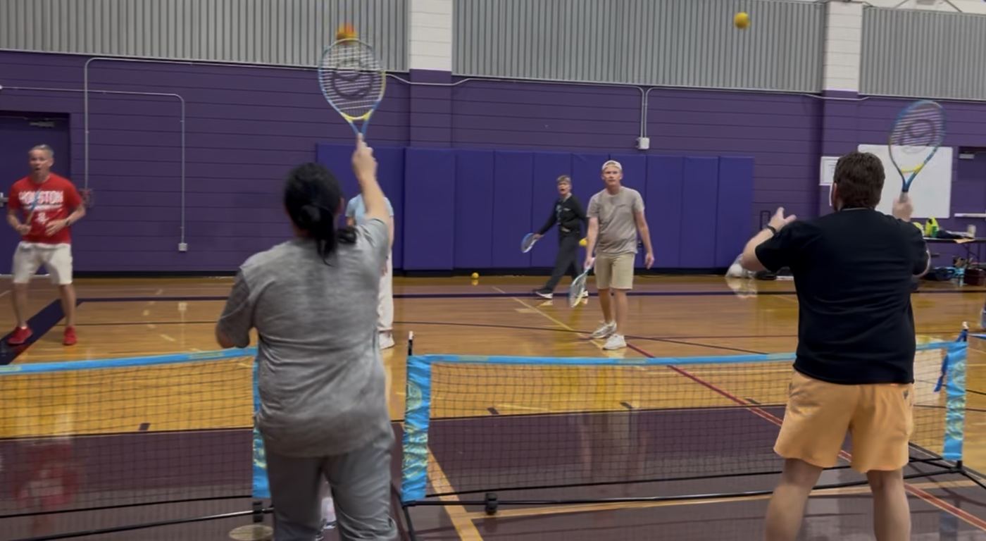 Adults rallying over a portable net on a shortened court, an orange ball in the air