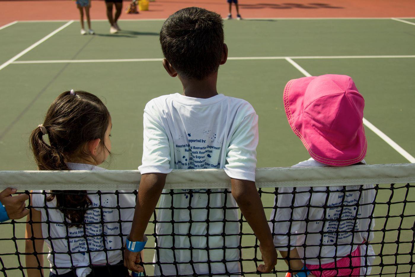 Three children watching from behind the net at a Houston NJTL session
